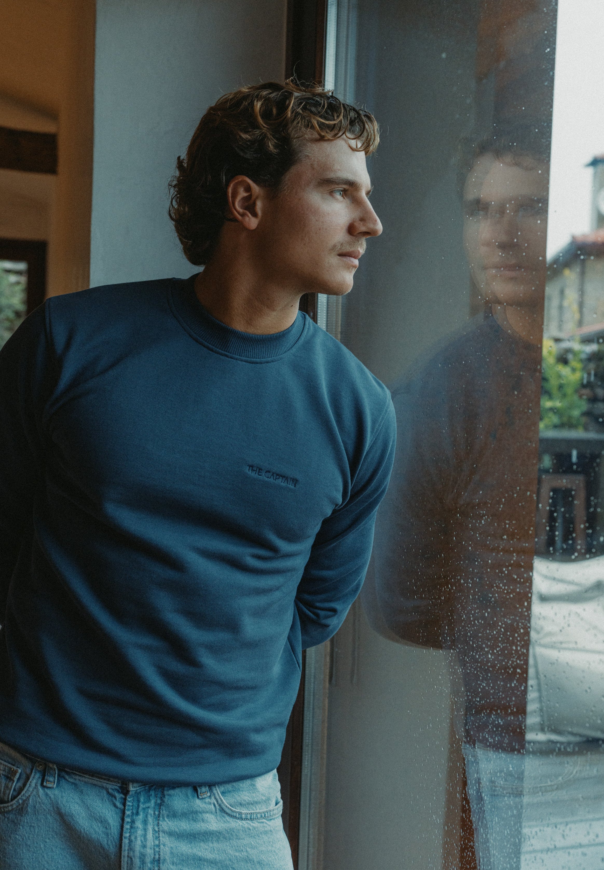 Man in a blue sweatshirt looking out of a window with a reflection.